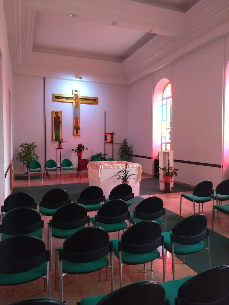 La cappella dell’Istituto: una sala bianca con un crocifisso dipinto e l’icona di sant’Ignazio dietro l’altare, una vetrata colorata e file di sedie