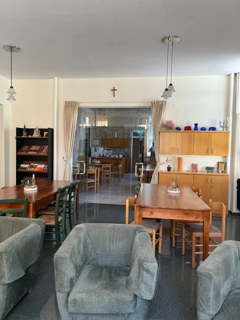 Una sala comune con poltrone grigie, tavoli e sedie di legno, una libreria e armadietti, e una parete di vetro che dà sulla sala da pranzo