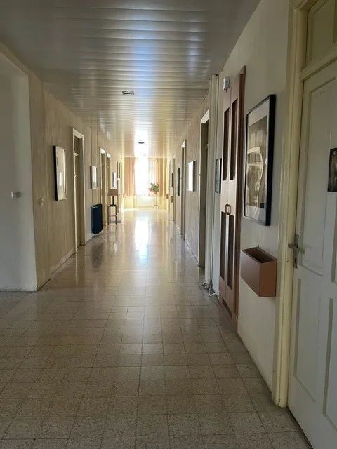 Un lungo corridoio con le porte delle camere ai due lati, quadri alle pareti e in fondo una finestra luminosa con tende