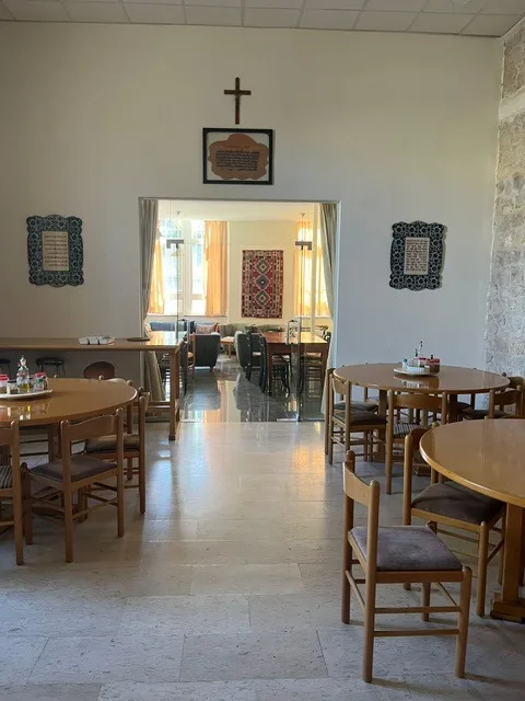 La sala da pranzo: tavoli rotondi e sedie di legno, piastrelle in ceramica e una croce alla parete, e una porta verso un salotto