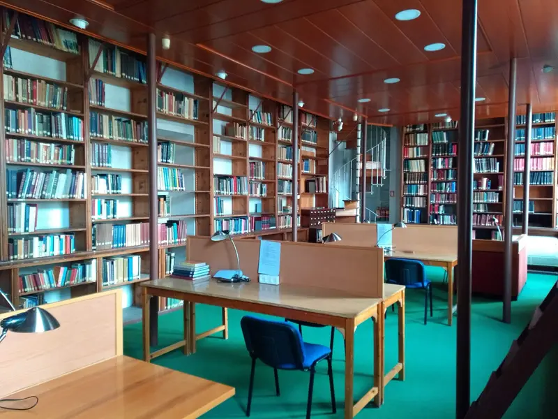 La sala di lettura della biblioteca: alte scaffalature in legno, scrivanie con lampade da lettura e sedie blu, e una scala a chiocciola verso i depositi